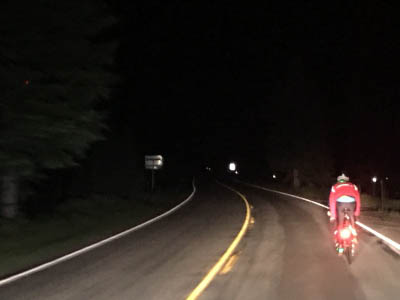 A tandem bicycle with a bright rear taillight and a cyclist wearing a red reflective jersey rides on smooth winding pavement partly lit up by the headlights of a vehicle behind.