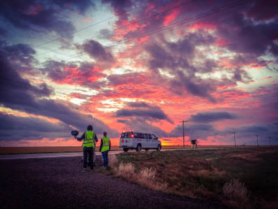 Two crew members in fluorescent yellow vests look on as a tandem bicycle rides away from a white support van. A pink and orange sunrise in Kansas lights up the background.